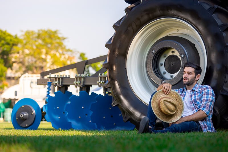 Agricultor sentado en llanta de tractor en campo rural al atardecer, maquinaria agrícola de fondo.